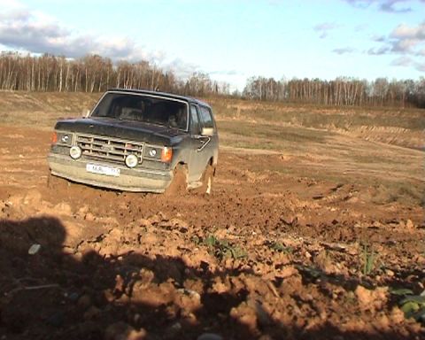 1987 Ford Bronco - A dark-colored Ford Bronco is stuck in thick mud with trees in the background under a partly cloudy sky.