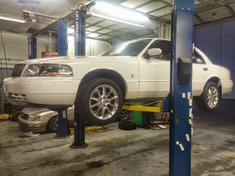 2000 Mercury Grand Marquis - A white 2000 Mercury Grand Marquis is raised on a car lift in a garage, with another vehicle visible underneath.