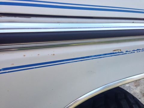 1979 Ford Bronco - Detail of a white vehicle's side panel blue racing stripes and chrome trim, with visible signs of rust.