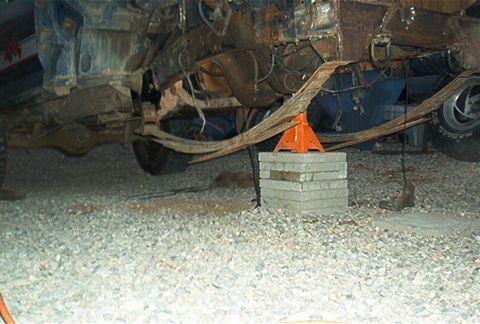 1986 Ford Bronco - Underbody view of a vehicle supported dangerously on a stack of concrete cinder blocks topped by an orange jack stand, with the leaf spring…