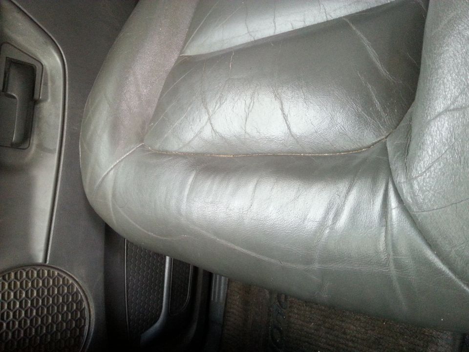 Close-up of a worn black leather driver's seat with visible creasing. The door panel and speaker grille are on the left
