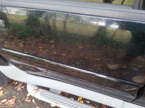 2001 Ford Explorer - Side view of a dark SUV door and running board, showing reflections of trees and fallen leaves on the ground.