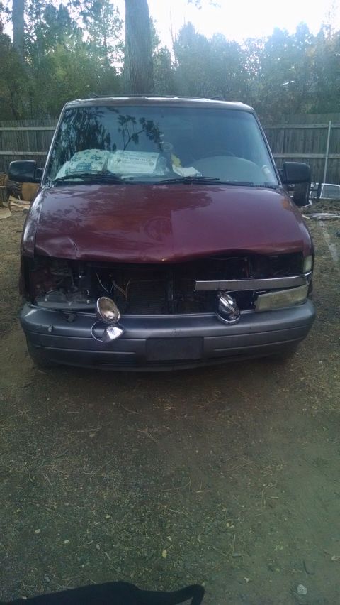 2000 Chevrolet Astro - The front of a damaged maroon van is shown, with a dented hood and missing headlights. The vehicle is parked on a gravel surface with a woo…