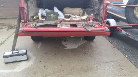 1981 Ford Bronco - The rear of a red pickup truck bed is open, showing various items including a toolbox, an ammo can, and camping gear. A trailer is attached…