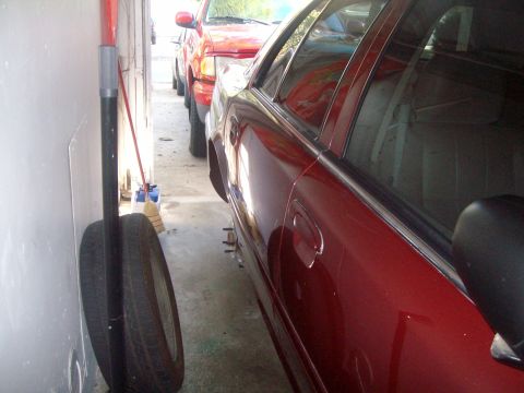 2004 Ford Crown Victoria - A red sedan is parked next to a red pickup truck in a narrow garage or driveway. A spare tire is leaning against a wall in the foreground.