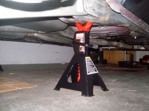 2004 Ford Crown Victoria - The underside of a vehicle is supported by a black jack stand with red accents. The exhaust system and fuel tank are visible.