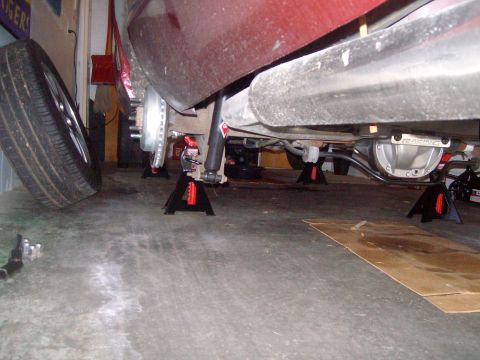 2004 Ford Crown Victoria - The underside of a red vehicle is supported by jack stands in a garage. The rear differential and exhaust system are visible, as well as th…