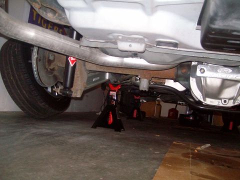 2004 Ford Crown Victoria - The underside of a Ford Crown Victoria is shown, supported by jack stands. Visible components include the exhaust system, rear differential…