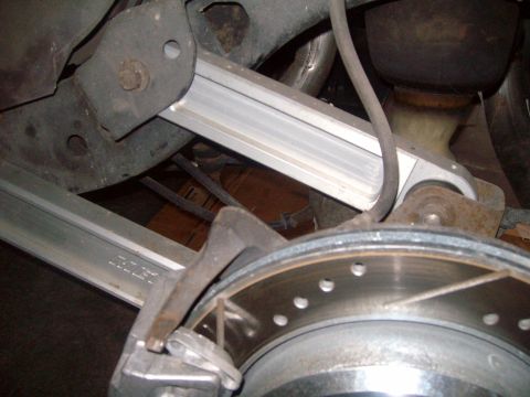2004 Ford Crown Victoria - A close-up view of the rear suspension of a vehicle, showing a silver aftermarket control arm installed near the brake rotor and caliper. T…