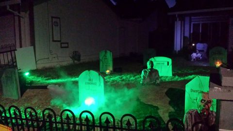 1989 Ford Bronco - A Halloween graveyard scene is set up in front of a house at night, complete with tombstones, fog, and zombie figures. Green lights illumin…
