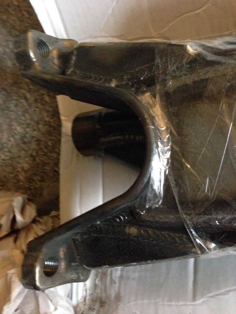 Black metal automotive suspension part with threaded holes, wrapped in plastic.
