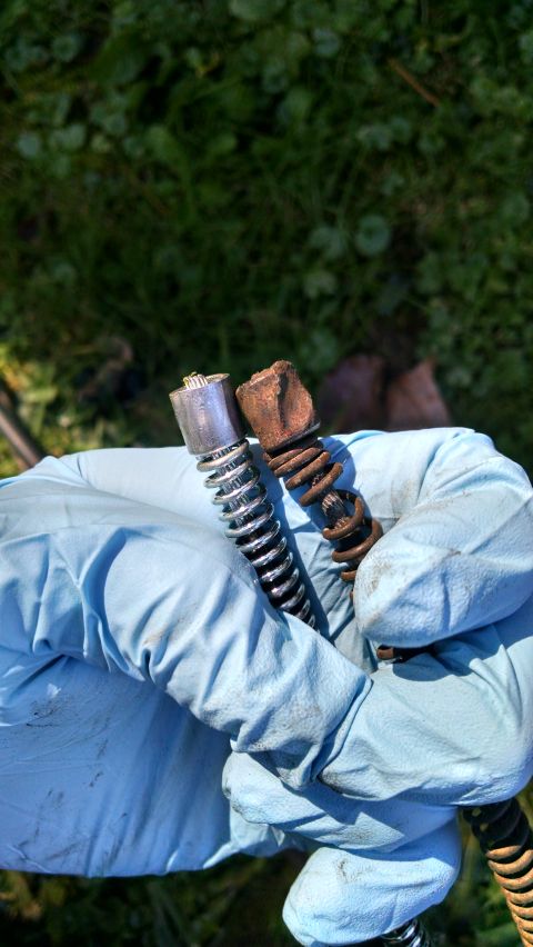 1994 Ford Bronco - A gloved hand holds two drain auger heads, one shiny and new, the other rusted and worn, against a blurred green background.