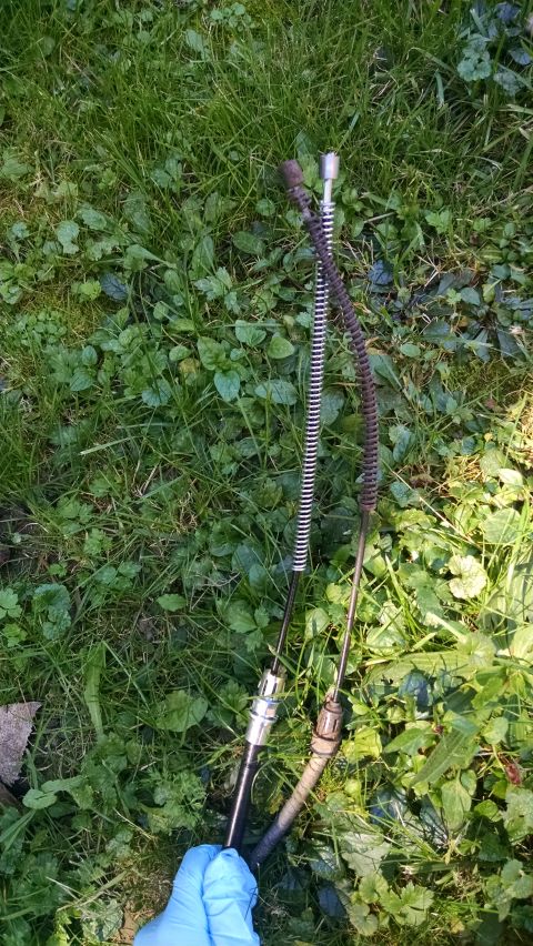 1994 Ford Bronco - Two brake cables are laid out on green grass. One cable has a silver spring and the other has a dark, coiled spring.