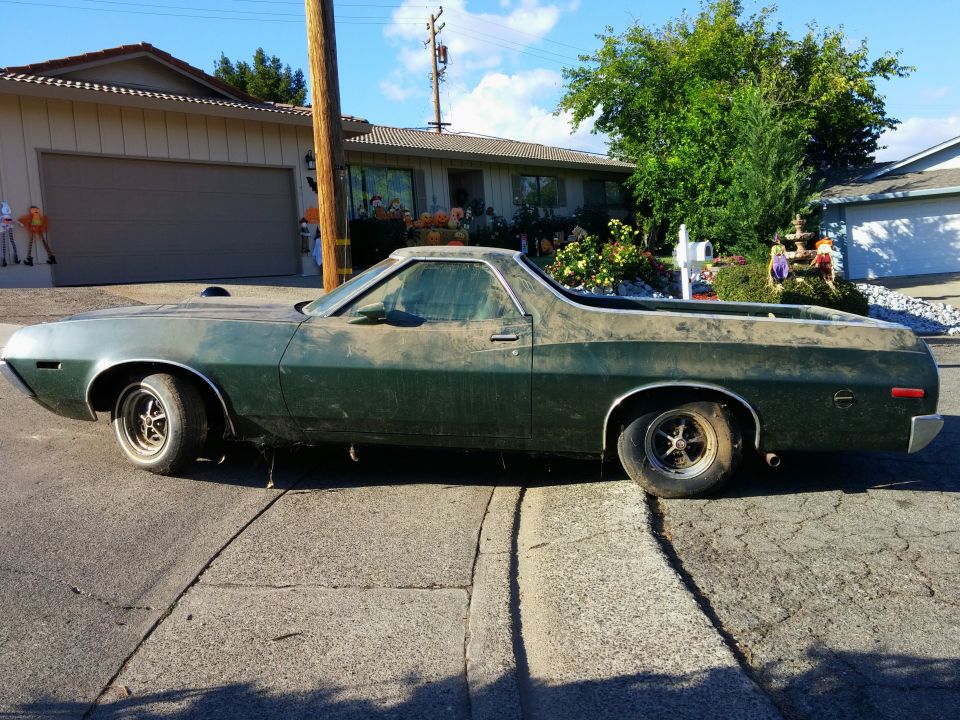 Dusty dark green 1970s Ford Ranchero with a flat tire, parked on a street with Halloween decorations.