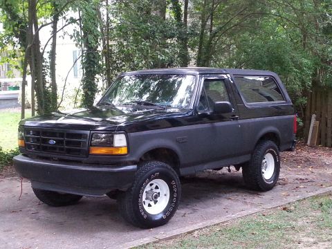 1994 Ford Bronco - A black 1994 Ford Bronco is parked on a concrete driveway surrounded by trees. The SUV has a two-tone paint scheme with a dark gray lower b…