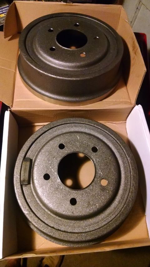 1994 Ford Bronco - Two new, dark gray brake drums are shown in an open cardboard box. The brake drums appear to be cast iron and are positioned one above the…