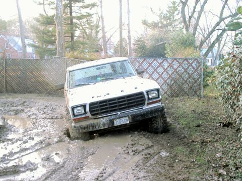 1978 Ford Bronco - A white 1978 Ford Bronco is stuck in deep mud, its front-left wheel submerged.