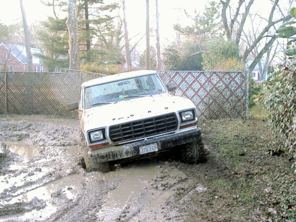 White 1978 Ford Bronco stuck in mud, front-left wheel deep, grille visible