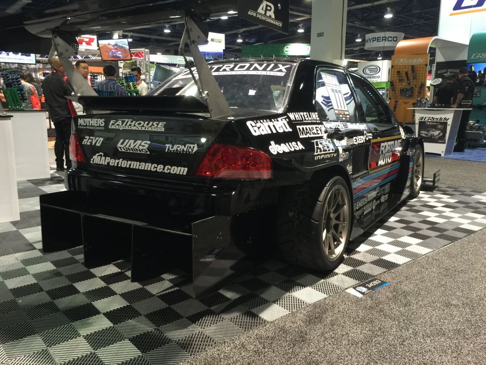 Modified black Mitsubishi Lancer Evolution with large wing and sponsor decals at an auto show.