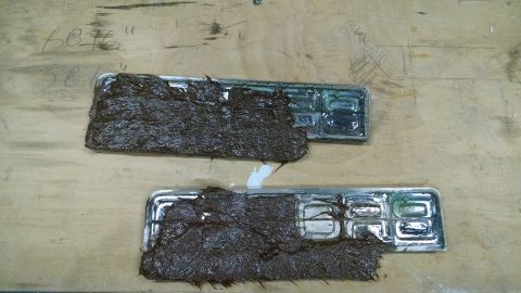 1992 Ford Bronco - Two chrome car emblems, partially obscured by mud, rest on a wooden surface with faint markings.