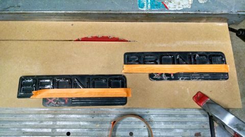 1992 Ford Bronco - Two 'Bronco' emblems are taped down on a workbench, with a table saw blade visible above them.