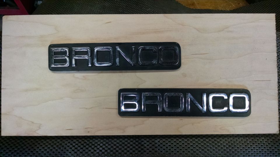 Two black Bronco emblems with chrome lettering on a light wood plank