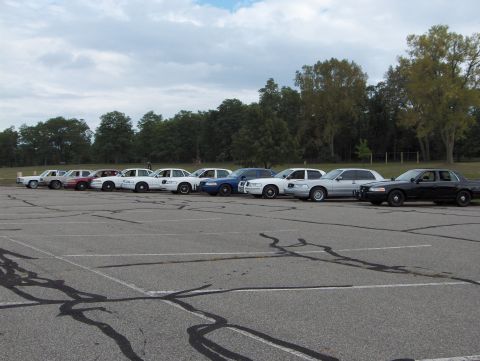 2007 Ford Crown Victoria - A lineup of Ford Crown Victorias and other sedans are parked in a lot under a cloudy sky.