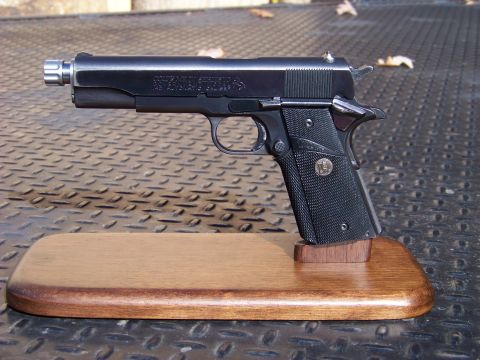 1995 Mercury Grand Marquis - A black Colt M1911 semi-automatic pistol with a silver muzzle attachment rests on a wooden display stand. The pistol is shown from the side…