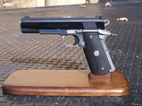 1995 Mercury Grand Marquis - A close-up, side view of a Colt MK IV Series 80 pistol displayed on a wooden stand. The pistol is black with silver accents and has texture…