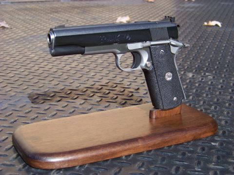 1995 Mercury Grand Marquis - A black and silver semi-automatic pistol is displayed on a wooden stand. The pistol is resting on a diamond plate surface with a few fallen…