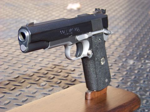 1995 Mercury Grand Marquis - A close-up, angled view of a black Colt MK IV Series 80 pistol resting on a wooden display stand. The pistol is shown from the front-left,…