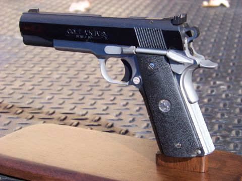 1995 Mercury Grand Marquis - A close-up, angled view of a black Colt MK IV Series 80 pistol resting on a wooden stand. The pistol features textured black grips with a s…