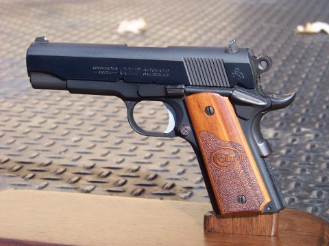 1995 Mercury Grand Marquis - A close-up, side profile of a black Colt Commander semi-automatic pistol with wooden grip panels. The pistol is resting on a wooden stand.