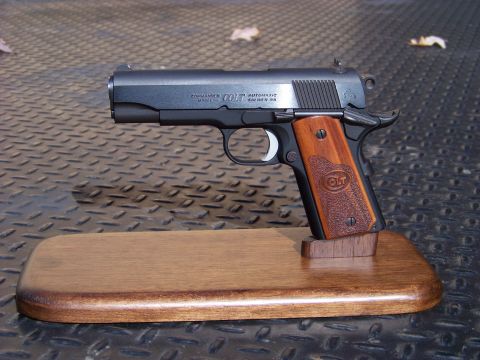 1995 Mercury Grand Marquis - A black Colt Commander automatic pistol chambered in .45 caliber rests on a wooden display stand. The pistol has wooden grips with the Colt…