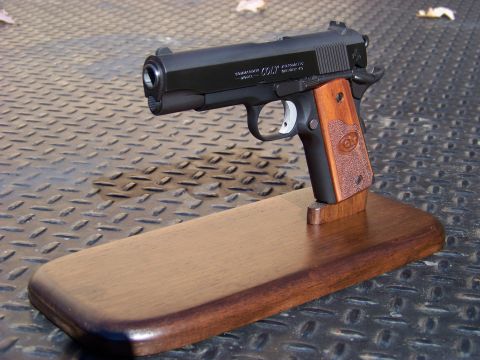 1995 Mercury Grand Marquis - A black Colt Commander semi-automatic pistol with wood grips is displayed on a wooden stand. The pistol is positioned on a textured metal s…