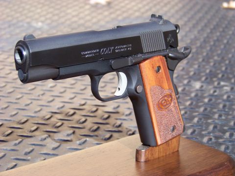 1995 Mercury Grand Marquis - A close-up, angled shot of a black Colt Commander semi-automatic pistol with wooden grips rests on a wooden stand. The pistol is displayed…