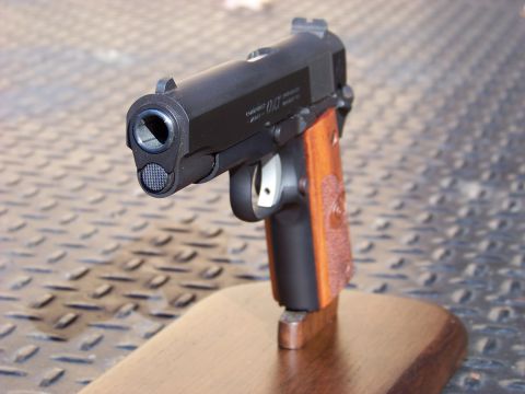 1995 Mercury Grand Marquis - A close-up, front-facing view of a black semi-automatic pistol with wooden grips, displayed on a wooden stand. The pistol is resting on a t…