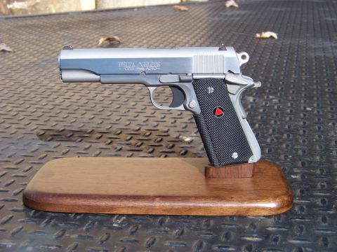 1995 Mercury Grand Marquis - A Colt Delta Elite 10mm pistol is displayed on a wooden stand. The pistol has a stainless steel finish and black checkered grips with a red…