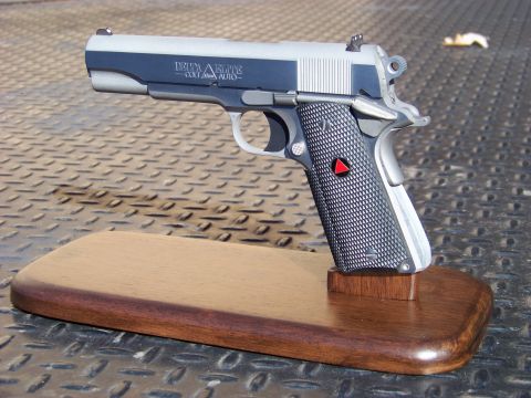 1995 Mercury Grand Marquis - A close-up view of a Colt Delta Elite 10mm Auto pistol displayed on a wooden stand. The pistol is silver and dark gray with black checkered…