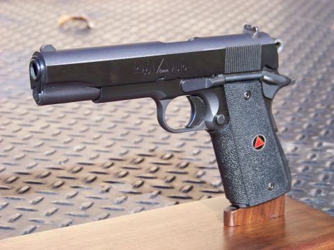 1995 Mercury Grand Marquis - A black Colt 1911 Delta Elite pistol is displayed on a wooden stand against a textured metal background. The pistol features textured grips…