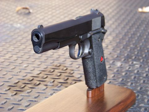 1995 Mercury Grand Marquis - A close-up, angled view of a black semi-automatic handgun with textured grips, resting on a wooden stand. The background is a textured meta…