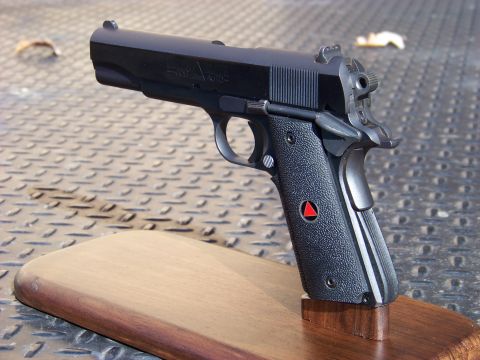 1995 Mercury Grand Marquis - A black Colt 1911 Delta Elite pistol is displayed on a wooden stand. The pistol has black textured grips with a red triangle emblem.