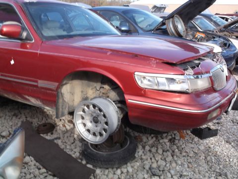1999 Mercury Grand Marquis - A red Mercury Grand Marquis is parked in a junkyard, missing a front wheel and with its hood ajar. Other vehicles and car parts are visible…