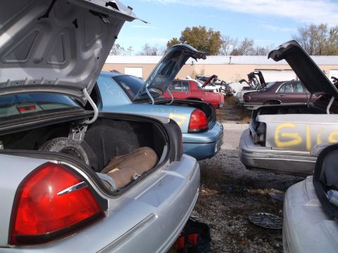 1999 Mercury Grand Marquis - A group of sedan vehicles with their trunks open are parked in a junkyard. One car in the foreground has a spare tire and a brown bag in it…