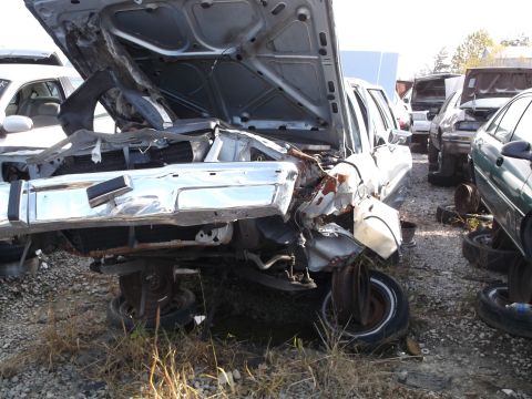 1999 Mercury Grand Marquis - A heavily damaged sedan with its hood open is parked in a junkyard. The front end is severely crumpled, revealing rusted metal and exposed…