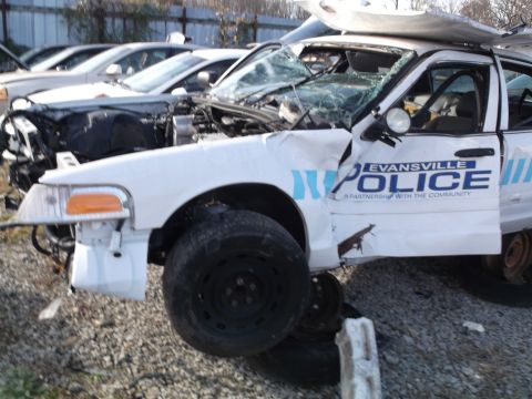 1999 Mercury Grand Marquis - A heavily damaged white police car, identified as an Evansville Police vehicle, is parked in a junkyard. The front end is crushed, the hood…