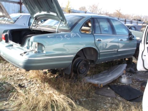 1999 Mercury Grand Marquis - A light blue sedan with its trunk open is parked in a junkyard. The car is missing its rear bumper and has one wheel visible.