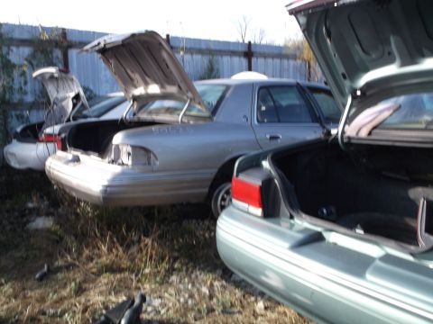 1999 Mercury Grand Marquis - Multiple sedans with their trunks open are parked in a junkyard setting. The closest vehicle is a light green sedan, while a silver sedan i…