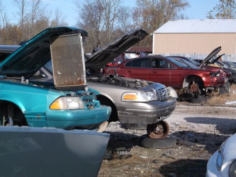 1999 Mercury Grand Marquis - A turquoise sedan in a junkyard has its hood open, revealing a large radiator. A red sedan is parked behind it, also with its hood open, an…