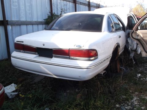 1999 Mercury Grand Marquis - The rear driver's side of a white sedan is shown parked in a grassy area next to a metal fence. The trunk lid has a Ford emblem, and the ta…
